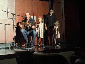man playing guitar with two little girls at microphones.