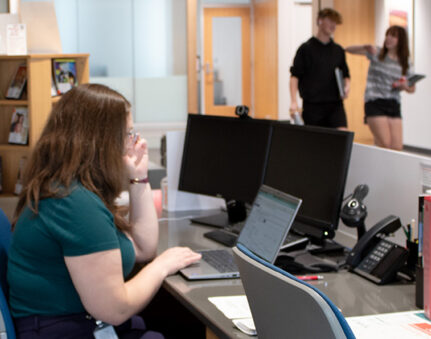 Liz Hartig on laptop with students in the background.
